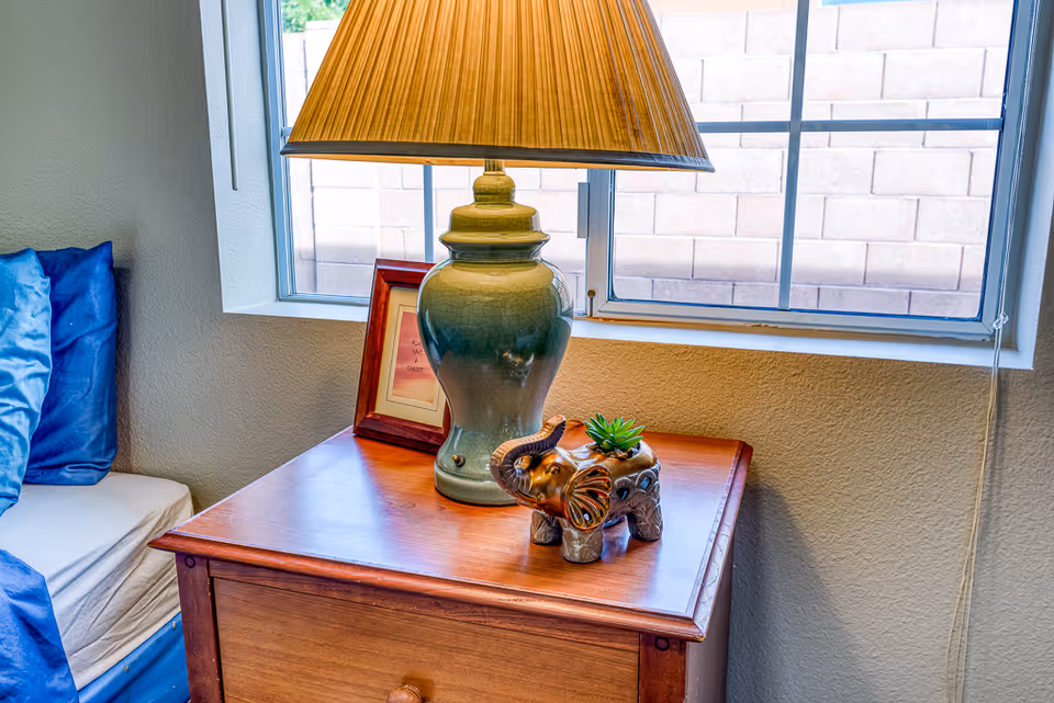 A wooden bedside table with a green ceramic lamp, a small framed picture, and a decorative elephant planter with a small succulent on top. The table is next to a bed with blue pillows and a window with a view of a brick wall behind it.