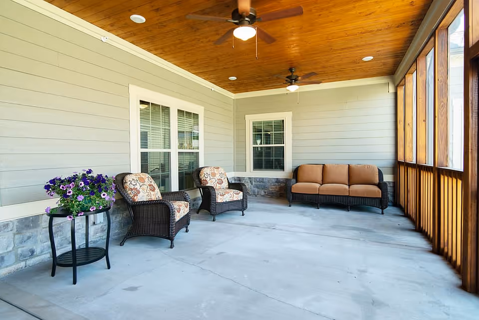 A screened-in porch area with a wooden ceiling featuring two ceiling fans. The porch has light gray walls with white trim and stone accents at the bottom. There are two cushioned wicker armchairs with floral patterned cushions, a cushioned wicker sofa with solid brown cushions, and a small black side table with a pot of purple flowers.