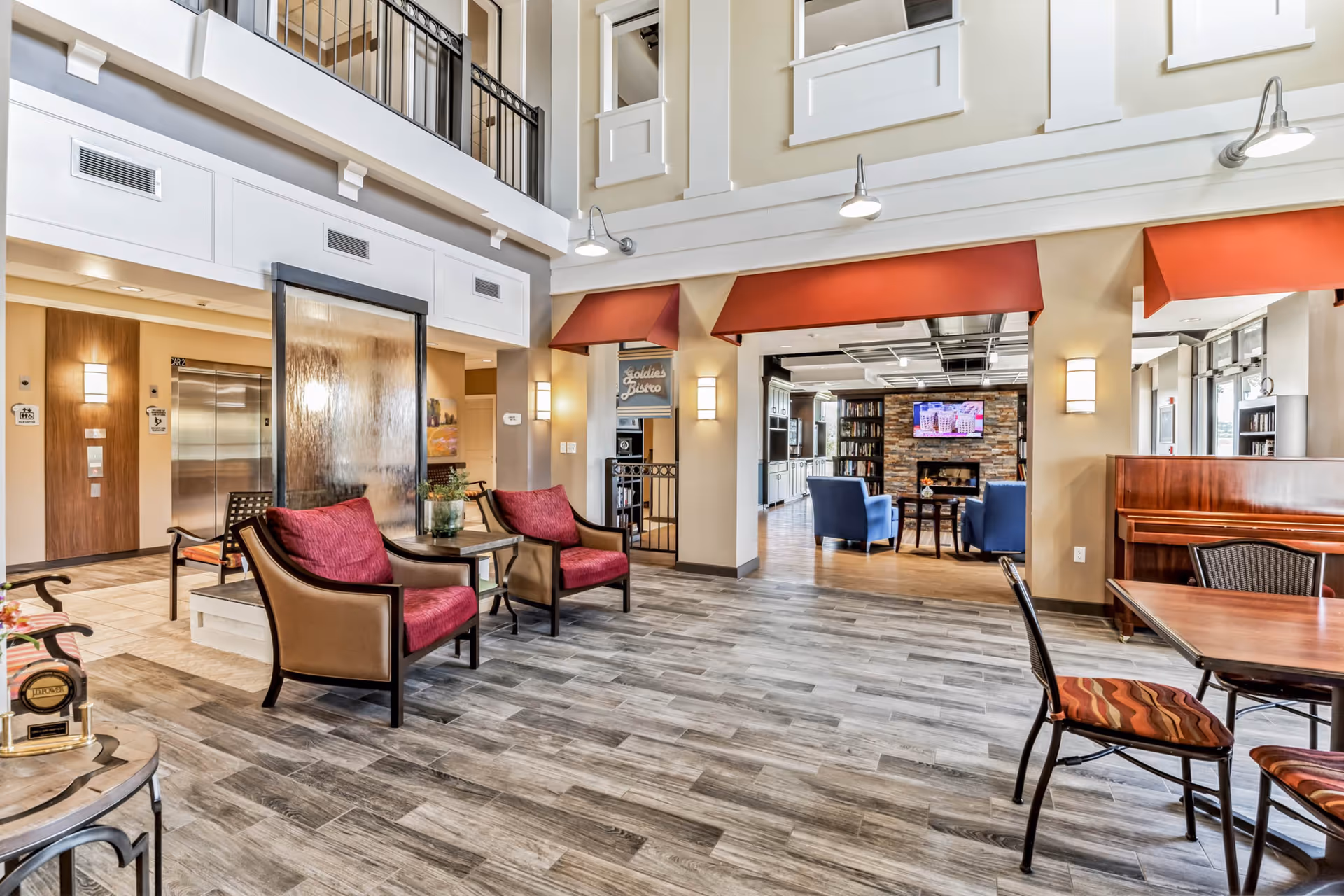 A spacious and well-lit common area in a senior living facility featuring comfortable seating with red cushions, a water feature, and a view into a cozy lounge with blue chairs and a fireplace. There is also a piano and dining table with chairs on the right side, and an elevator area with restrooms visible in the background.