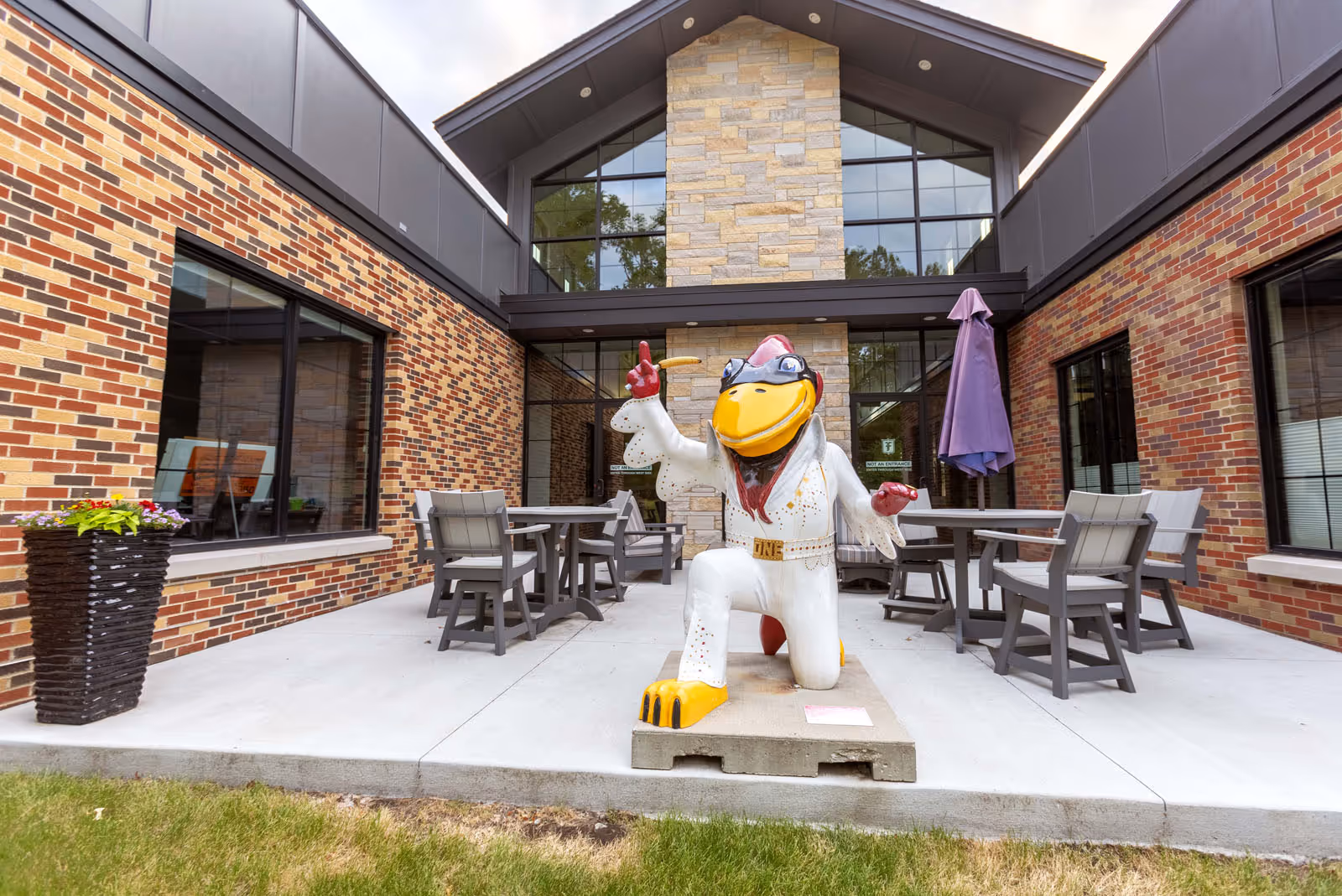 Outdoor courtyard patio with tables and chairs and a large colorful bird statue in front of a brick building.