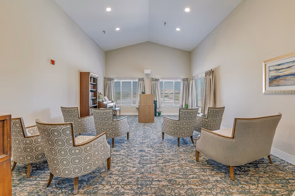 Bright communal seating area with patterned armchairs arranged facing a podium and windows in a senior living facility.