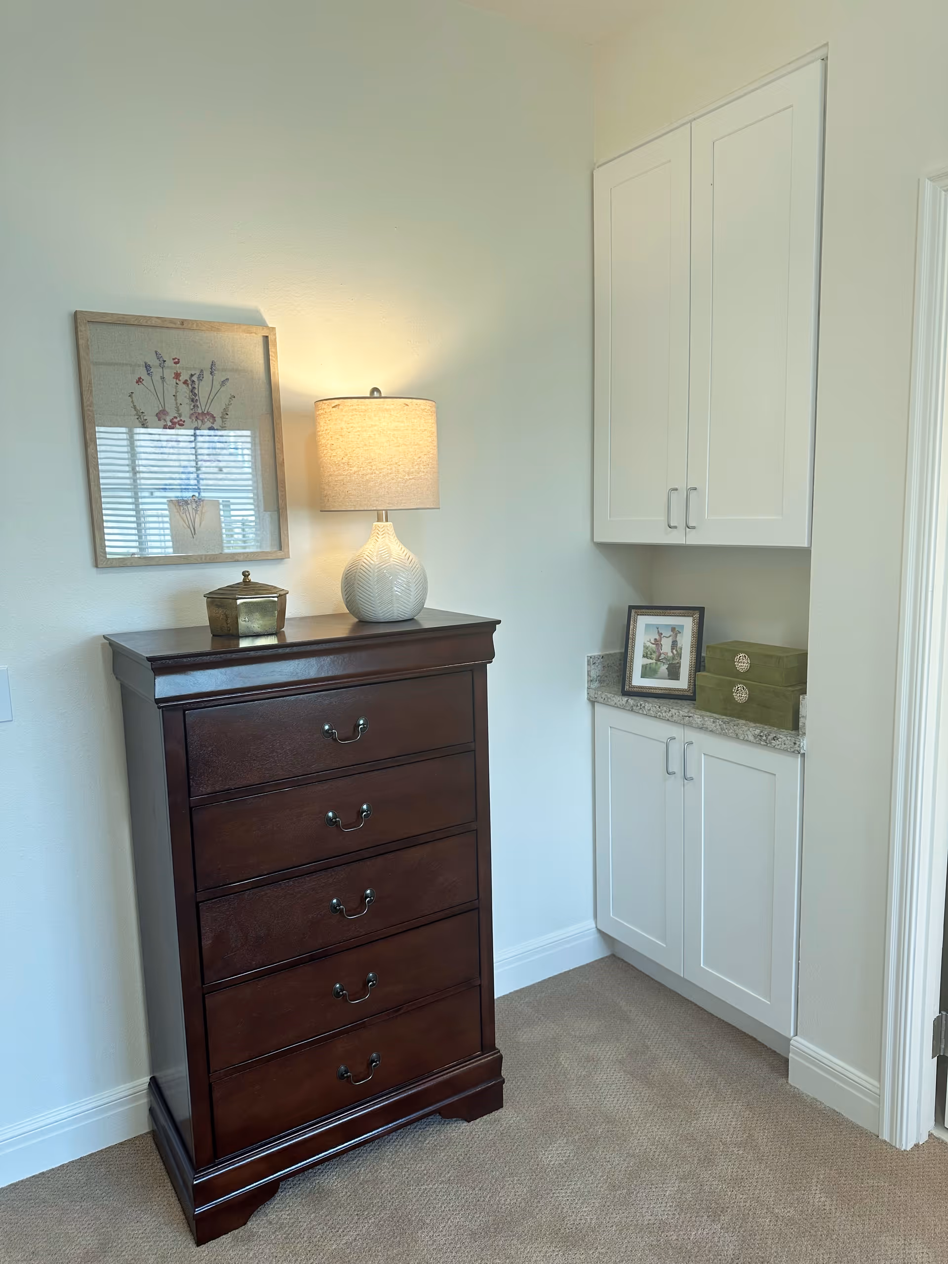A corner of a room featuring a dark wooden chest of drawers with five drawers, topped with a white textured table lamp and a small decorative box. Above the chest is a framed picture of flowers. To the right, there is a built-in white cabinet with upper and lower doors, a granite countertop with a framed photo and two green decorative boxes. The room has beige carpet and light-colored walls.