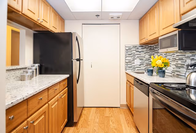 A modern kitchen with wooden cabinets, granite countertops, a black refrigerator, a stainless steel dishwasher, a microwave, an electric stove with a kettle, and a backsplash with a mosaic tile design. There is a vase with yellow flowers on the counter.