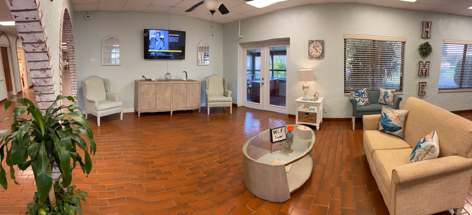 A cozy living room area with a beige sofa adorned with ocean-themed pillows, a glass-top oval coffee table with a Wi-Fi sign and a small flower vase, two light green armchairs, a wooden cabinet with decorative items, a wall-mounted TV, a side table with a lamp, and windows with blinds. The room has brick flooring, light blue walls, and a ceiling fan. There is a large green plant near a white brick archway leading to another area.