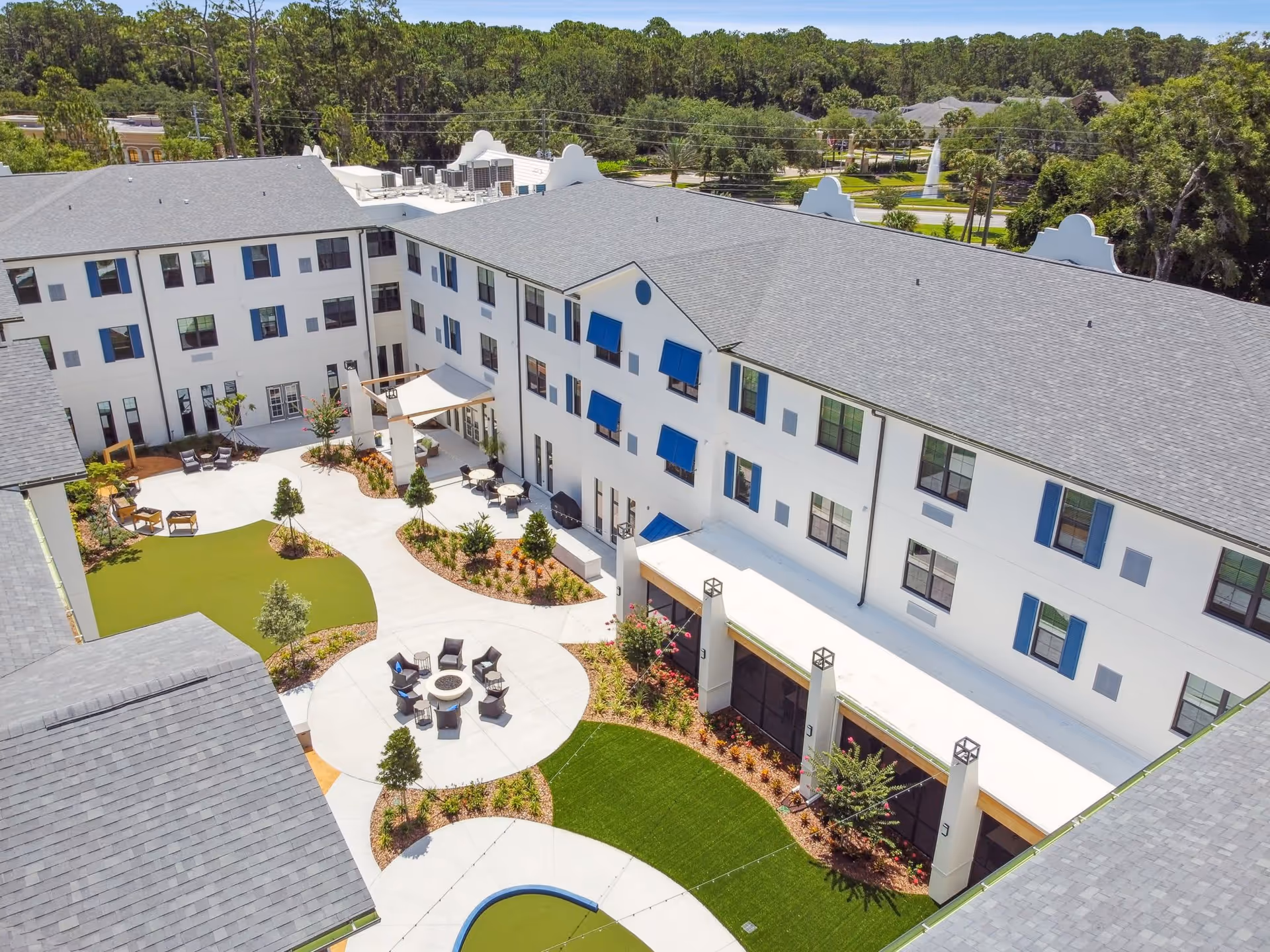 Aerial view of an outdoor courtyard area at Aden Senior Living featuring a circular seating area with chairs around a fire pit, several tables with chairs under a canopy, landscaped garden beds, and a well-maintained lawn surrounded by a three-story white building with blue window awnings.