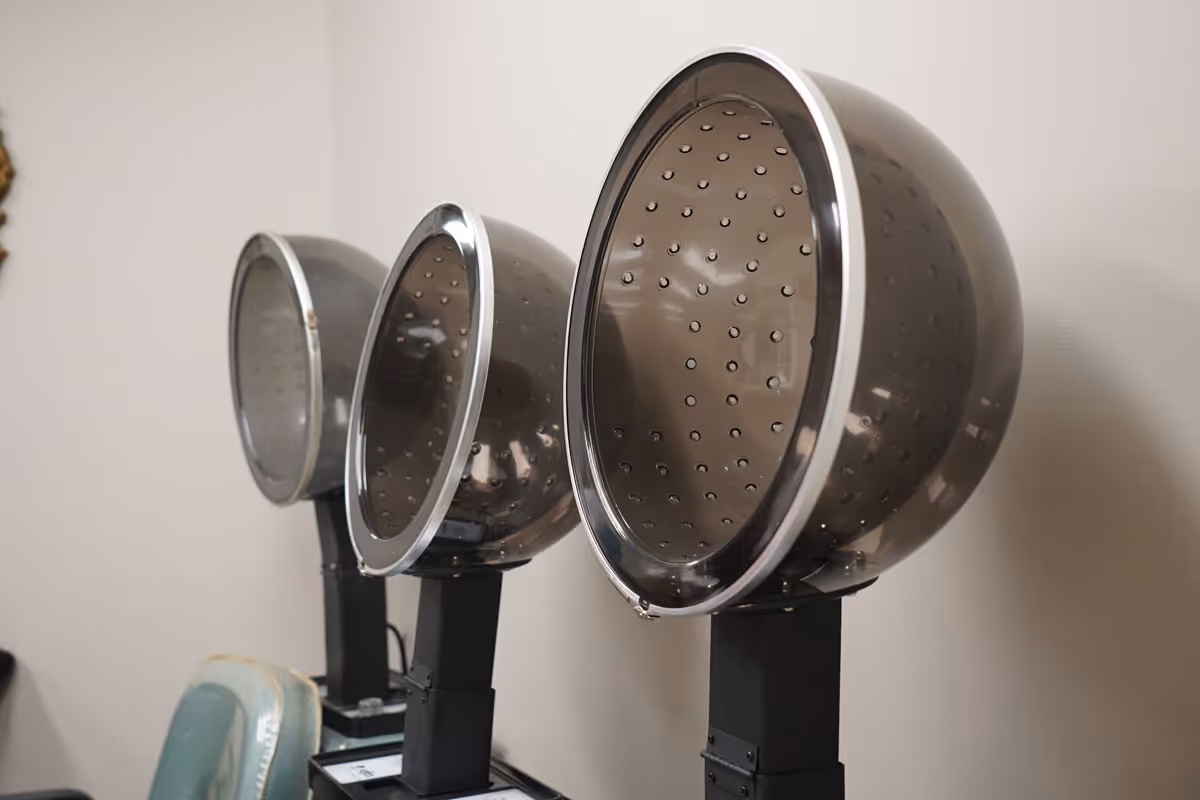 Three vintage hair dryer hoods mounted on stands in a salon or beauty area with a light-colored wall background.
