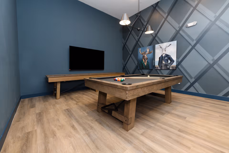 A modern game room with a wooden pool table in the center, a wall-mounted flat-screen TV, and two paintings of animals dressed in suits on a dark blue wall with a geometric pattern. The room has light wood flooring and a contemporary ceiling light fixture.