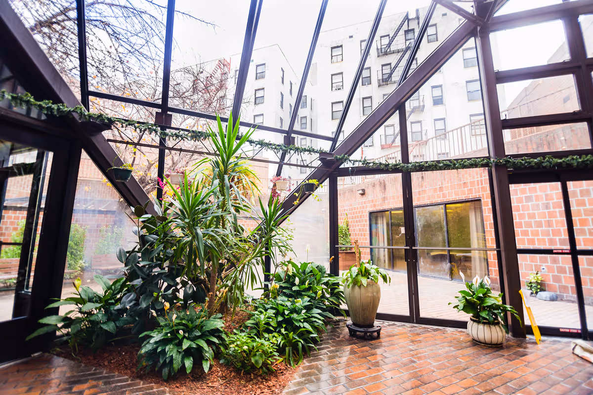 Indoor garden area with various green plants and potted plants inside a glass-enclosed space with brick flooring and a view of adjacent buildings through the glass walls.