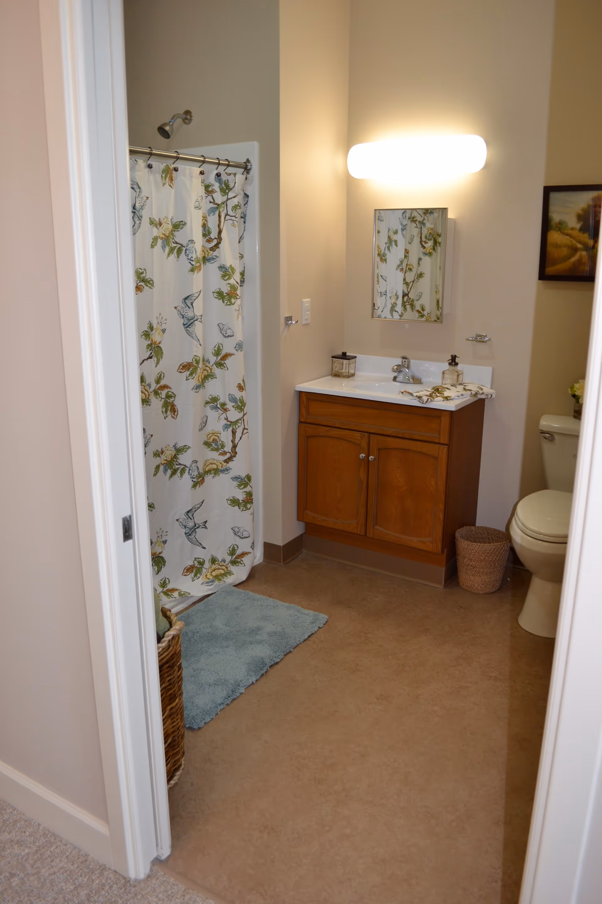 A bathroom with a shower area featuring a floral and bird patterned shower curtain, a wooden vanity with a white countertop and sink, a mirror above the sink, a wall-mounted light fixture, a toilet, a small waste basket, a blue bath mat on the floor, and a framed picture on the wall.