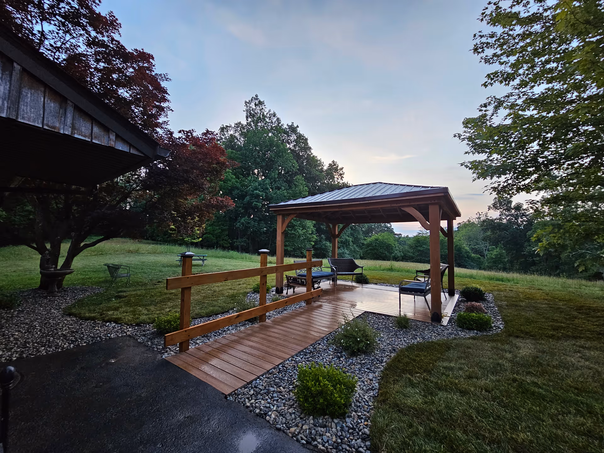 A wooden gazebo with a metal roof situated in a grassy outdoor area surrounded by trees. The gazebo has seating including chairs and a bench, and a wooden ramp leading up to it. The surrounding landscape includes a lawn, bushes, and a tree with red leaves on the left side.