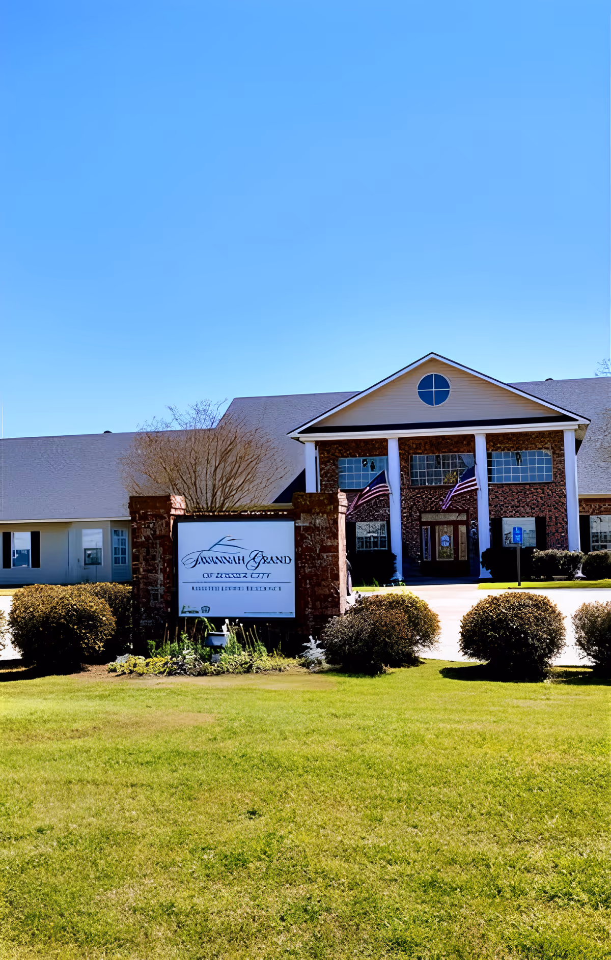 Front exterior of a senior living facility showing the building entrance with columns, American flags, a lawn, and a sign reading Savannah Grand of Bossier City.