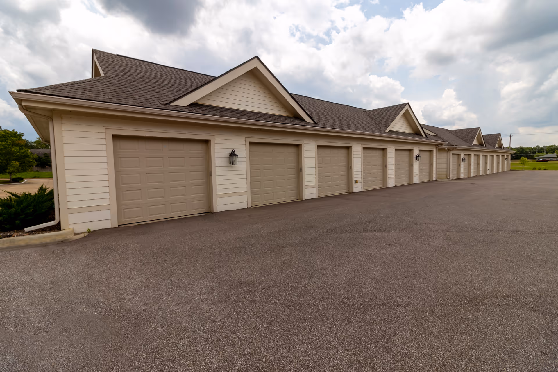 A long row of beige garage doors attached to a building with a sloped roof under a cloudy sky, with a large paved area in front.