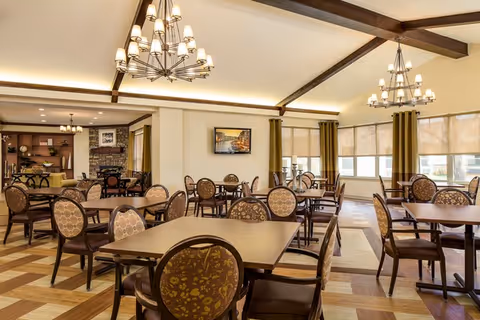 A spacious dining room with multiple wooden tables and chairs featuring floral upholstery. The room has large windows with beige roller shades and green curtains, wooden ceiling beams, and two large chandeliers. A flat-screen TV is mounted on the wall, and there is a cozy seating area with a fireplace in the background.