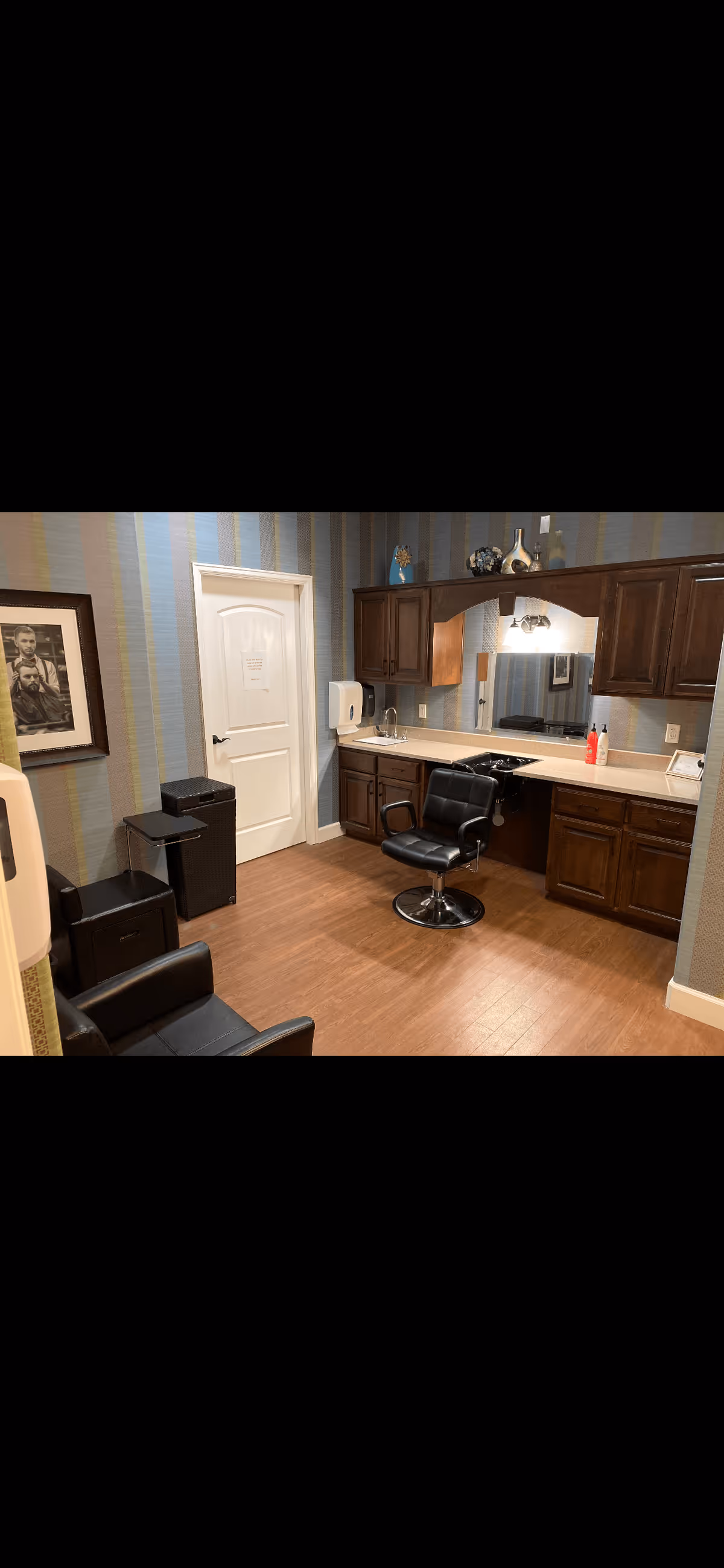 A small hair salon or barber station with a black salon chair in front of a countertop with a sink. The countertop is surrounded by wooden cabinets and a large mirror. The room has striped wallpaper in shades of blue, green, and beige, and a wooden floor. There is a white door, a framed black and white photo on the wall, and some decorative items on top of the cabinets.