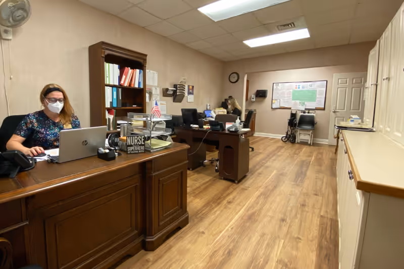 An office space with two women working at desks with laptops. One woman is wearing a face mask and sitting at a wooden desk with a sign that reads 'NURSE SUPERHERO'. The room has wooden flooring, beige walls, a bookshelf with binders, and cabinets along one wall. There is a clock on the wall and a wheelchair near the back.