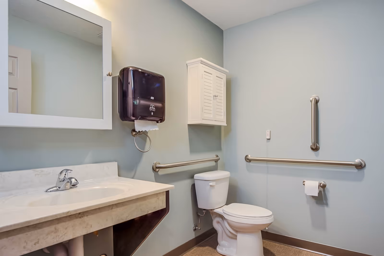 Accessible bathroom with a sink and mirror, toilet, grab bars, wall cabinet, and paper dispenser.