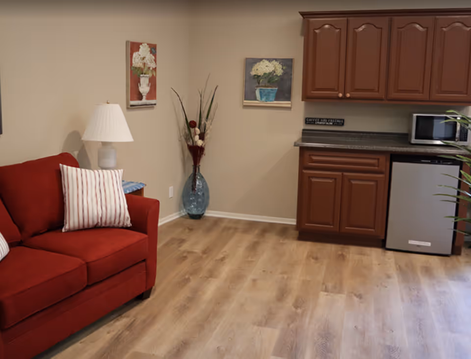 Interior view of a living area in Villa Sorrento Assisted Living featuring a red couch with striped pillows, a white table lamp on a small side table, two framed floral paintings on the beige wall, a tall decorative vase with dried flowers, wooden cabinets with a countertop, a microwave, and a small refrigerator, all set on a light wood floor.