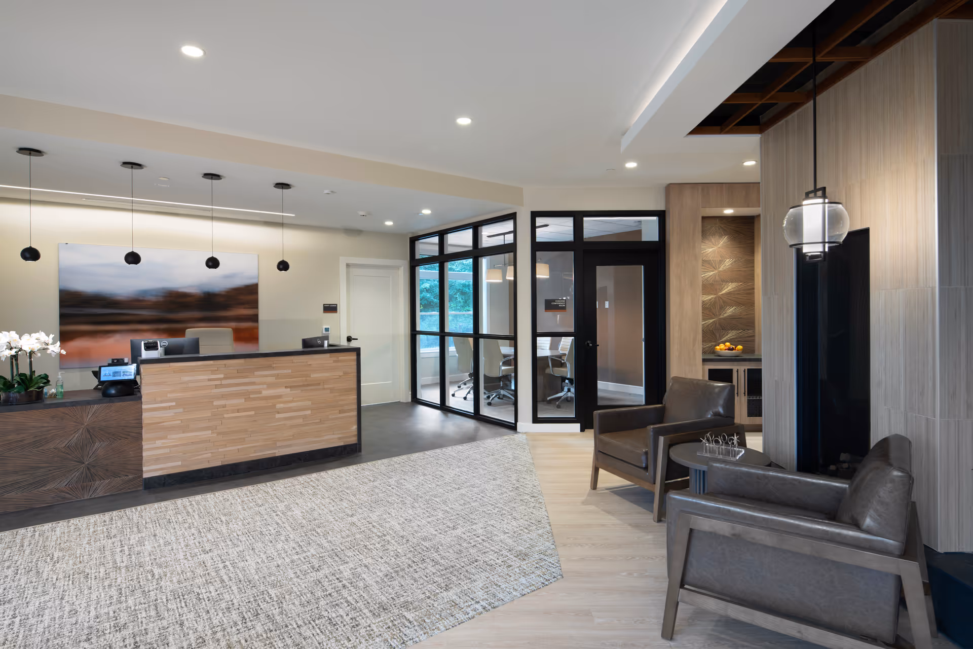 Modern reception lobby with a wood-paneled front desk, leather seating area, and glass-walled office in the background.