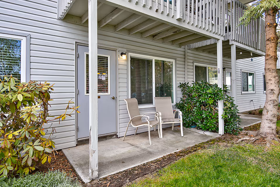 Covered ground-level patio with two outdoor chairs beside a door and windows on a light-gray sided building, bordered by shrubs and lawn.
