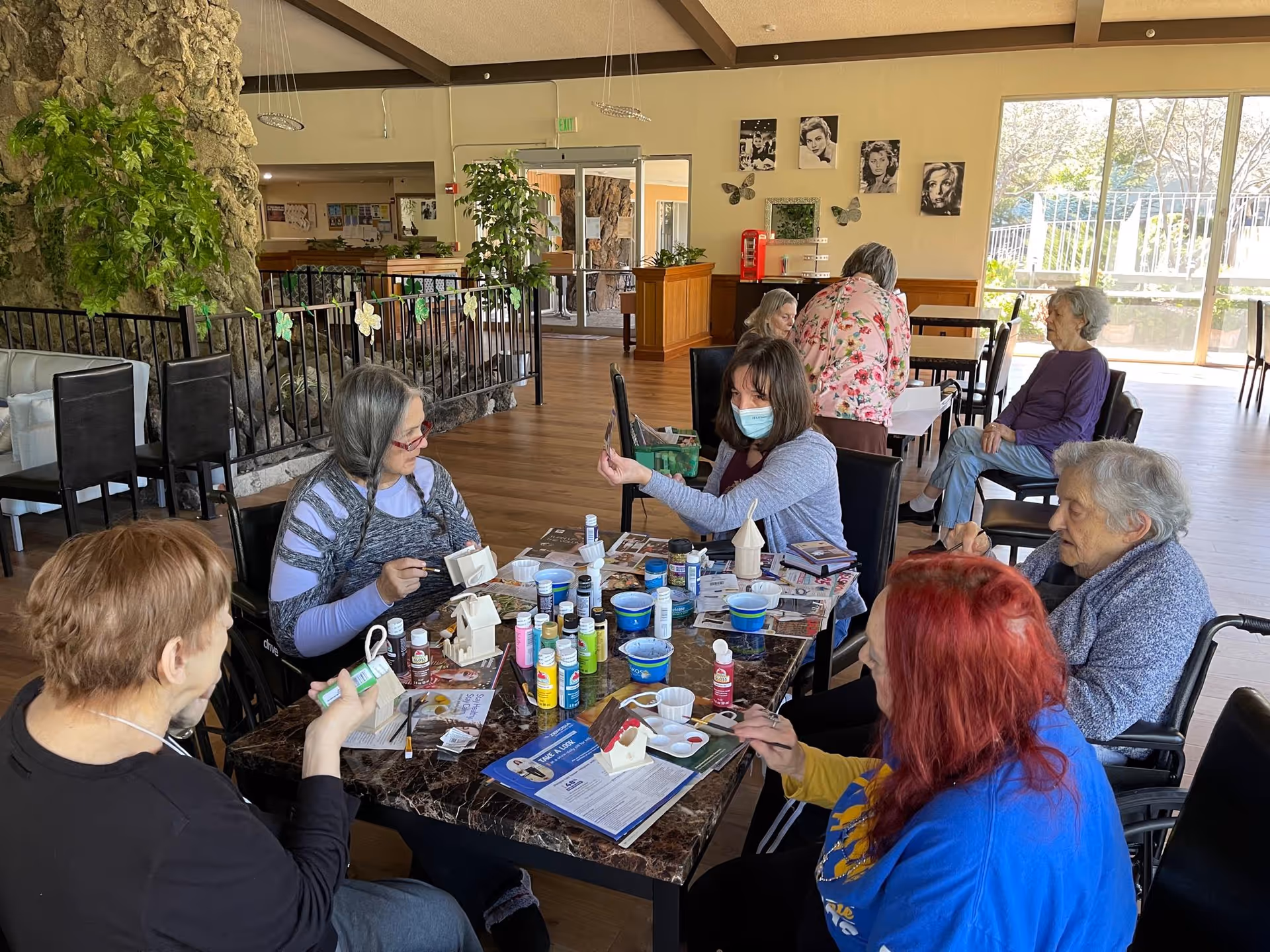 A group of elderly residents and a staff member wearing a mask gathered around a table painting crafts in a bright communal room.