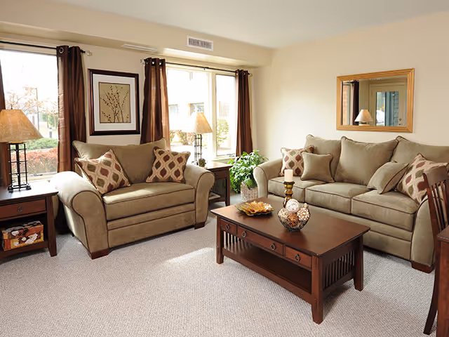 Sunlit living room with two beige sofas, a wooden coffee table, side tables with lamps, and decorative pillows in front of large windows.