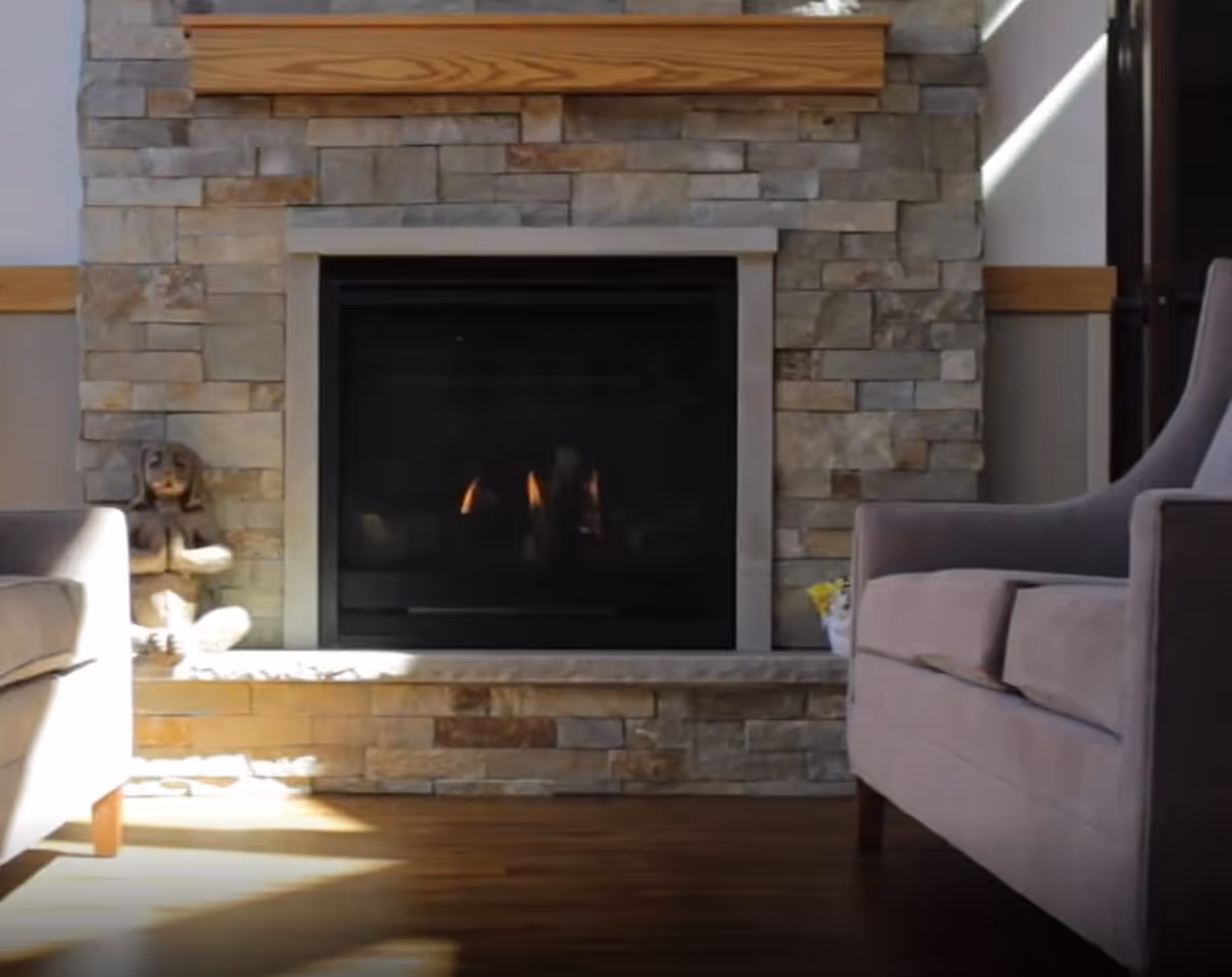 A cozy living room area featuring a stone fireplace with a wooden mantle. There is a small statue of a meditating figure on the left side of the fireplace and a small potted plant with yellow flowers on the right side. Two gray upholstered armchairs are partially visible on either side of the fireplace. Sunlight streams in, casting shadows on the wooden floor.