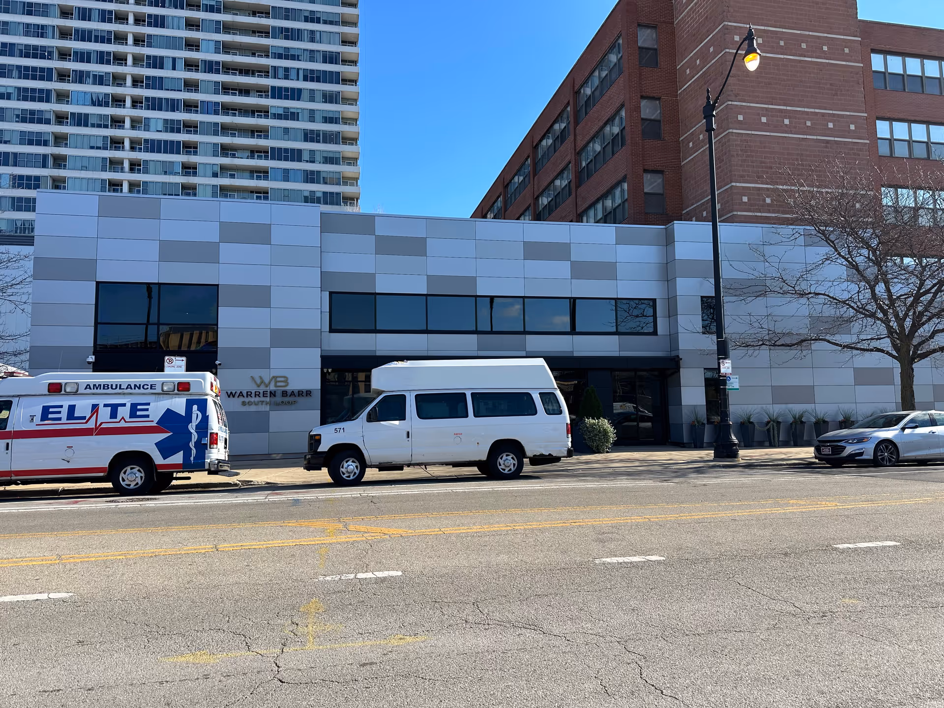 Street view of the Warren Barr South Loop facility building with a modern gray and white exterior. There are three vehicles parked in front: an ambulance with 'ELITE' written on the side, a white van, and a silver car. A tall residential building and a brick building are visible in the background under a clear blue sky.