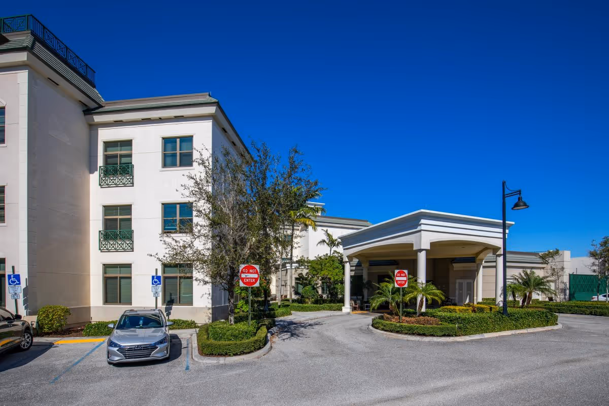 Entrance driveway and covered portico of a multi-story senior living building with parked cars and landscaping under a clear blue sky.