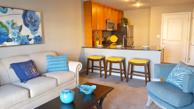 A cozy living room area with a beige sofa adorned with blue pillows, a blue armchair with a patterned pillow, and a dark wooden coffee table holding blue decorative items. In the background, there is a kitchen with wooden cabinets, stainless steel appliances, and a counter with three yellow cushioned stools. A large floral painting with blue flowers hangs on the wall above the sofa.
