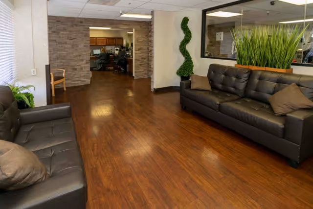 Interior view of a waiting area with two black leather sofas facing each other, a wooden floor, a tall green plant in a planter, and an open doorway leading to an office area with desks and computers.