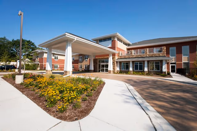 Exterior view of a modern senior living facility with a covered entrance, landscaped flower beds with yellow flowers, and a clear blue sky.