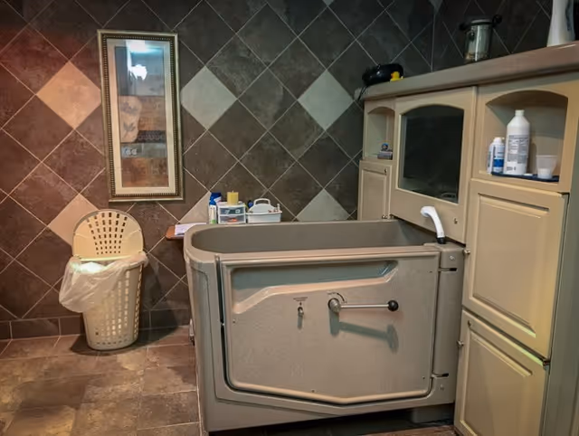 A bathing area in a senior living facility featuring a walk-in bathtub with a door and handle, beige cabinetry with various bottles and containers on the countertop, a laundry basket with a plastic liner, and a framed decorative picture on a tiled wall.