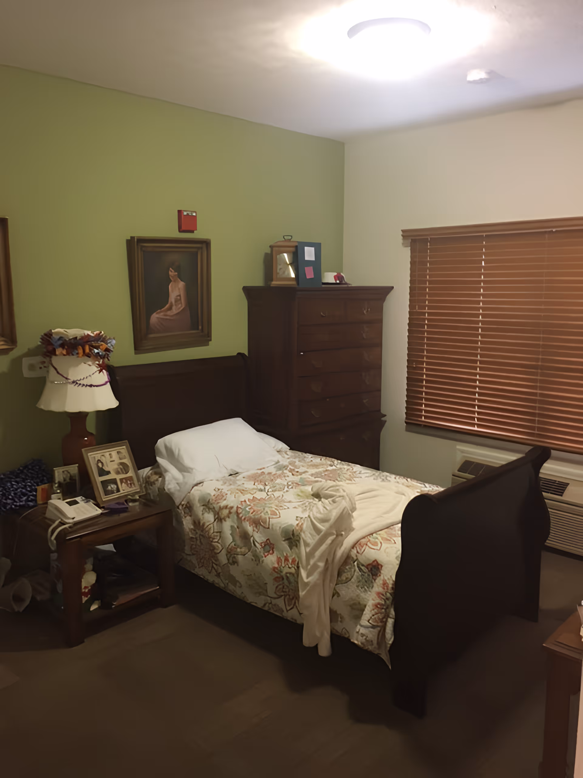 A cozy bedroom with a single wooden bed covered with a floral bedspread and a white pillow. A cream-colored blanket is draped over the bed. Next to the bed is a wooden nightstand with a lamp, framed photos, and a telephone. Behind the bed is a green accent wall with a framed painting of a woman. A tall wooden dresser stands against the adjacent wall. The window has brown blinds, and there is an air conditioning unit below it. The ceiling light is on, illuminating the room.