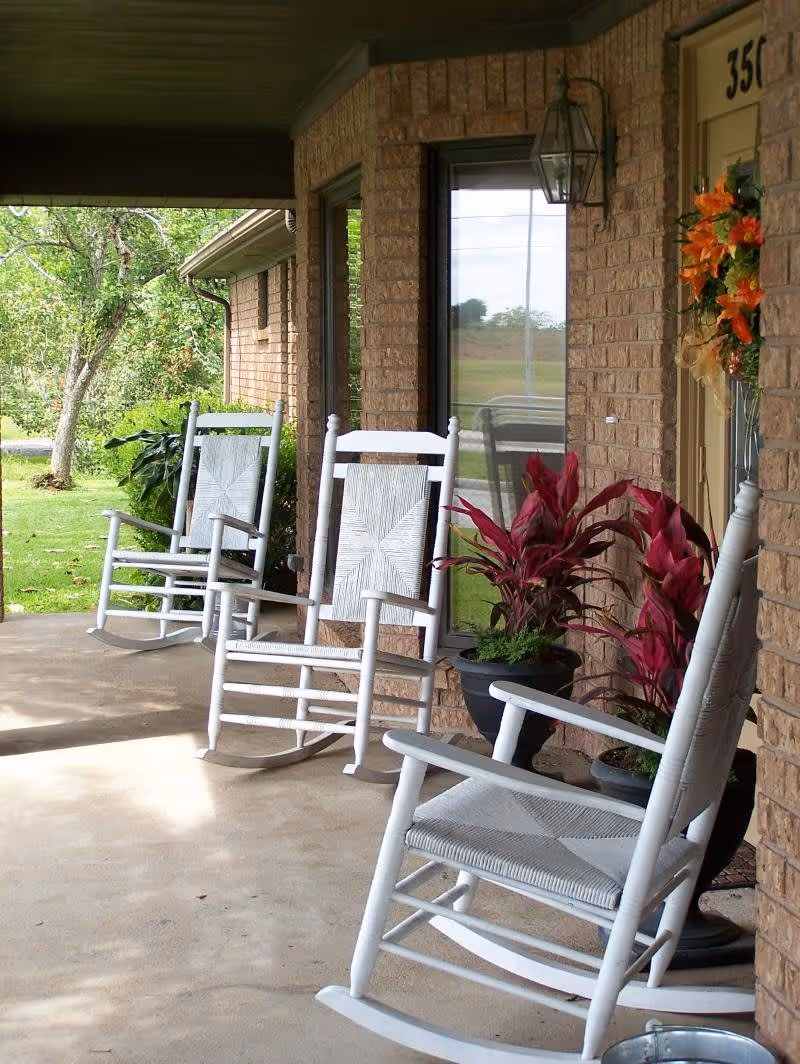 A covered porch area with three white wooden rocking chairs arranged along a brick wall. There are potted plants with red leaves placed near the chairs, and a door decorated with an orange floral wreath. Green trees and grass are visible in the background.