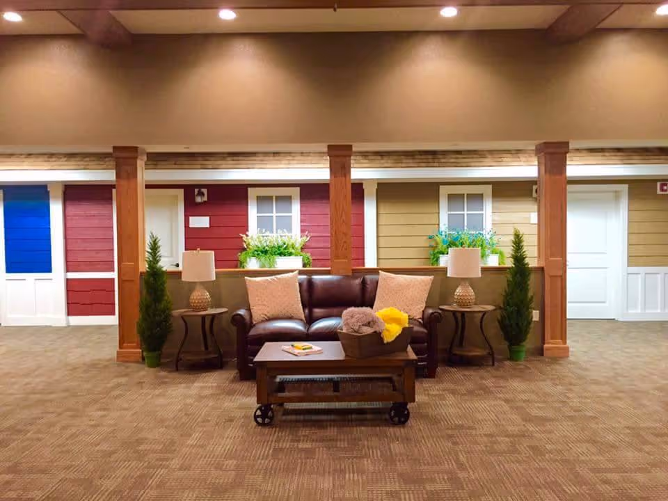 Indoor common area designed to resemble a small neighborhood street with colorful house facades, including blue, red, and beige walls with windows and doors. In the foreground, there is a brown leather sofa with two beige pillows, flanked by two side tables each with a lamp and a small potted plant. A coffee table with a basket containing blankets and a book is placed in front of the sofa. The area is carpeted and has wooden beams on the ceiling.