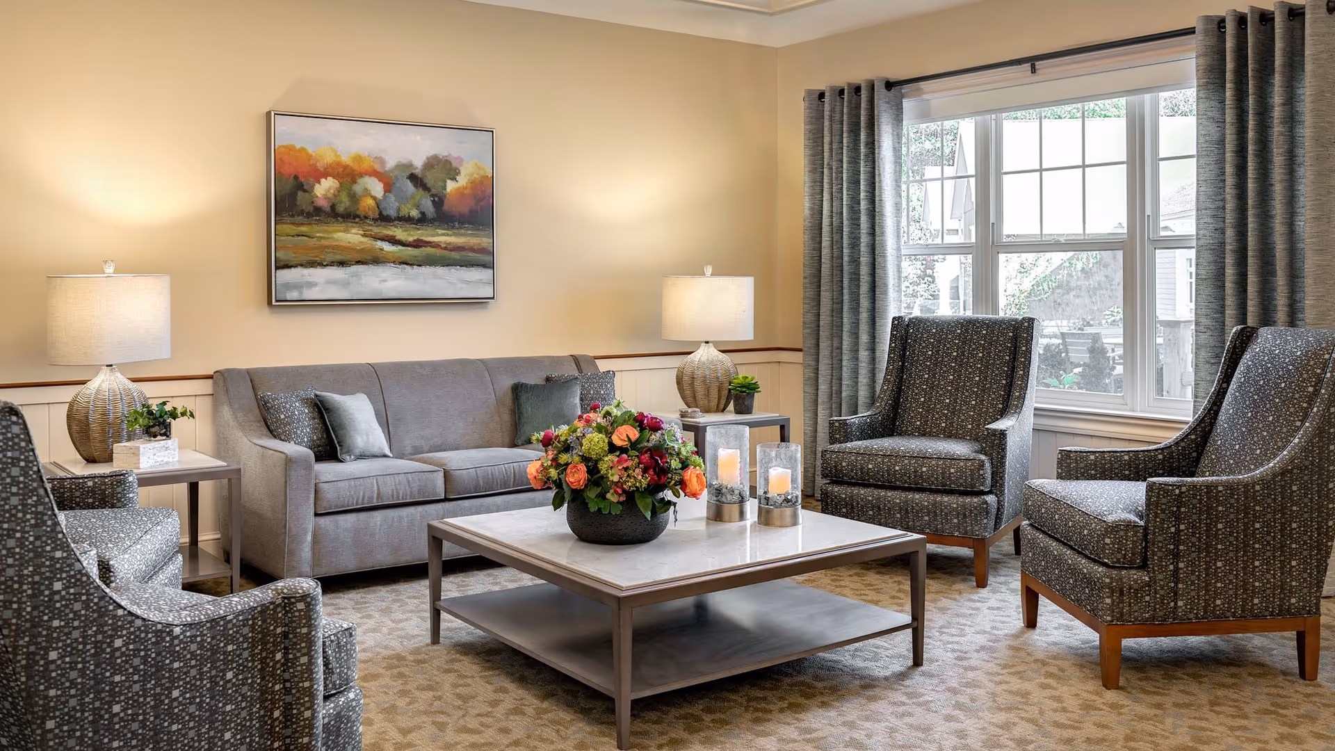 Well-lit living room with a sofa, four armchairs, a central coffee table with a floral arrangement, table lamps, artwork, and a large window.