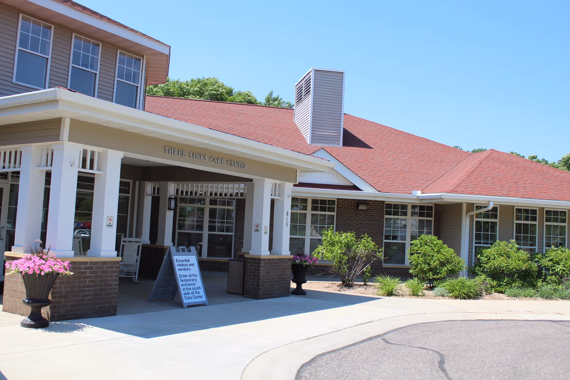 Exterior view of Three Links Care Center building with a covered entrance, brick and siding walls, multiple windows, and a red roof. There are flower pots with pink flowers and green shrubs around the entrance area under a clear blue sky.