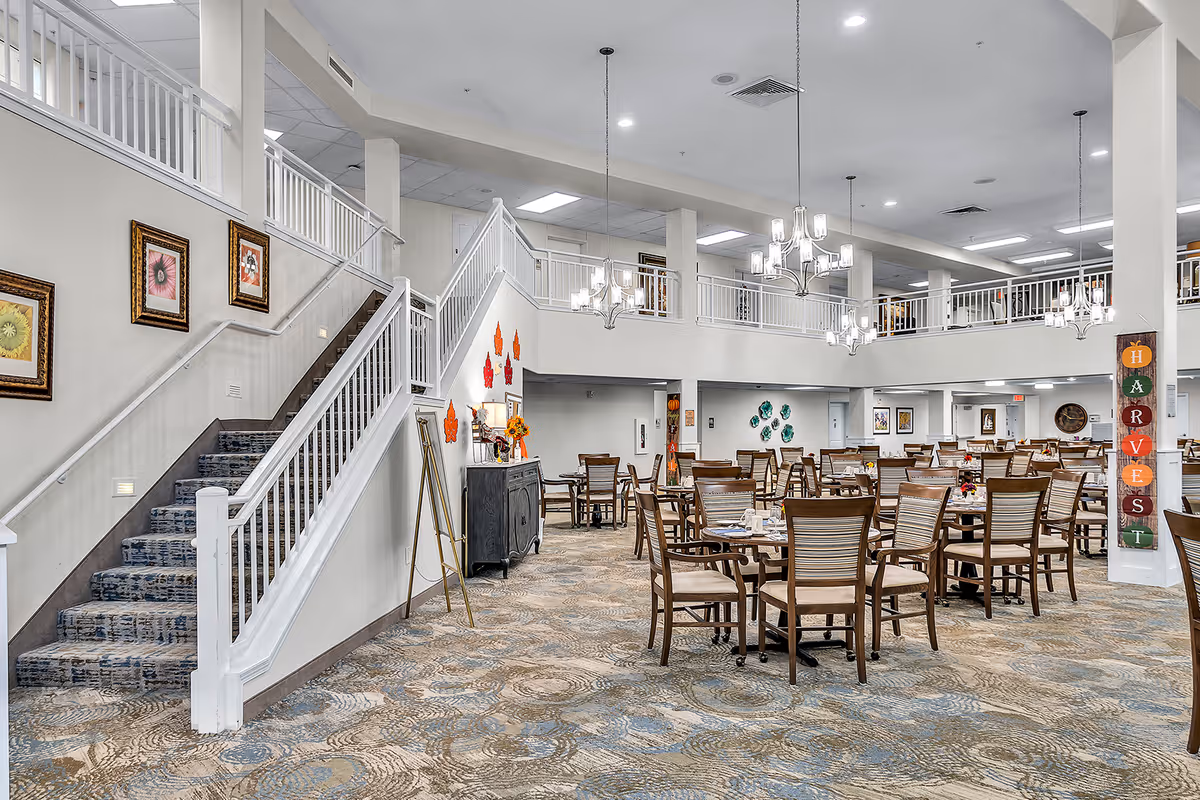 Spacious dining area in a retirement living facility with multiple wooden tables and chairs arranged neatly. The room features a high ceiling with chandeliers, a staircase with white railings leading to an upper level, and decorative wall art including framed pictures and colorful leaf decorations. The carpet has a patterned design, and the overall ambiance is bright and welcoming.