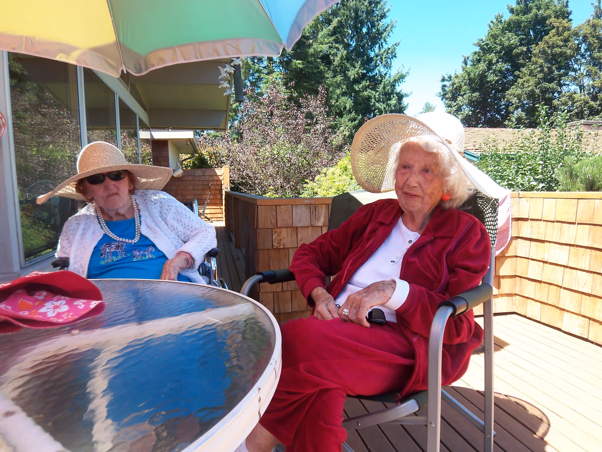 Two elderly women sitting outside on a wooden deck under a large umbrella. One woman is wearing a large straw hat, sunglasses, a white cardigan, and a blue shirt with a zebra design. The other woman is wearing a large white sunhat, a red jacket, and red pants. There are trees and bushes in the background under a clear blue sky.