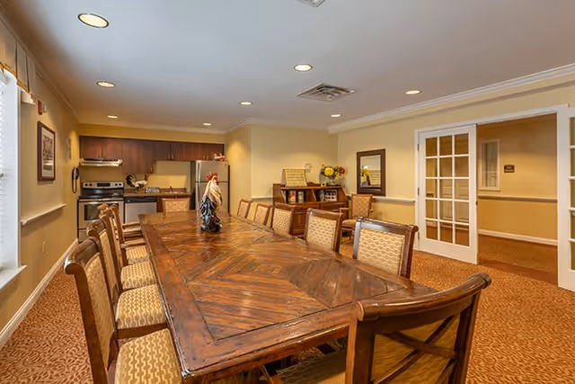A spacious dining room with a long wooden table surrounded by cushioned chairs. The room has warm lighting with recessed ceiling lights, a kitchen area with stainless steel appliances including a stove and refrigerator, and a sideboard with decorative items and flowers. There are windows with curtains on the left and a set of French doors on the right.