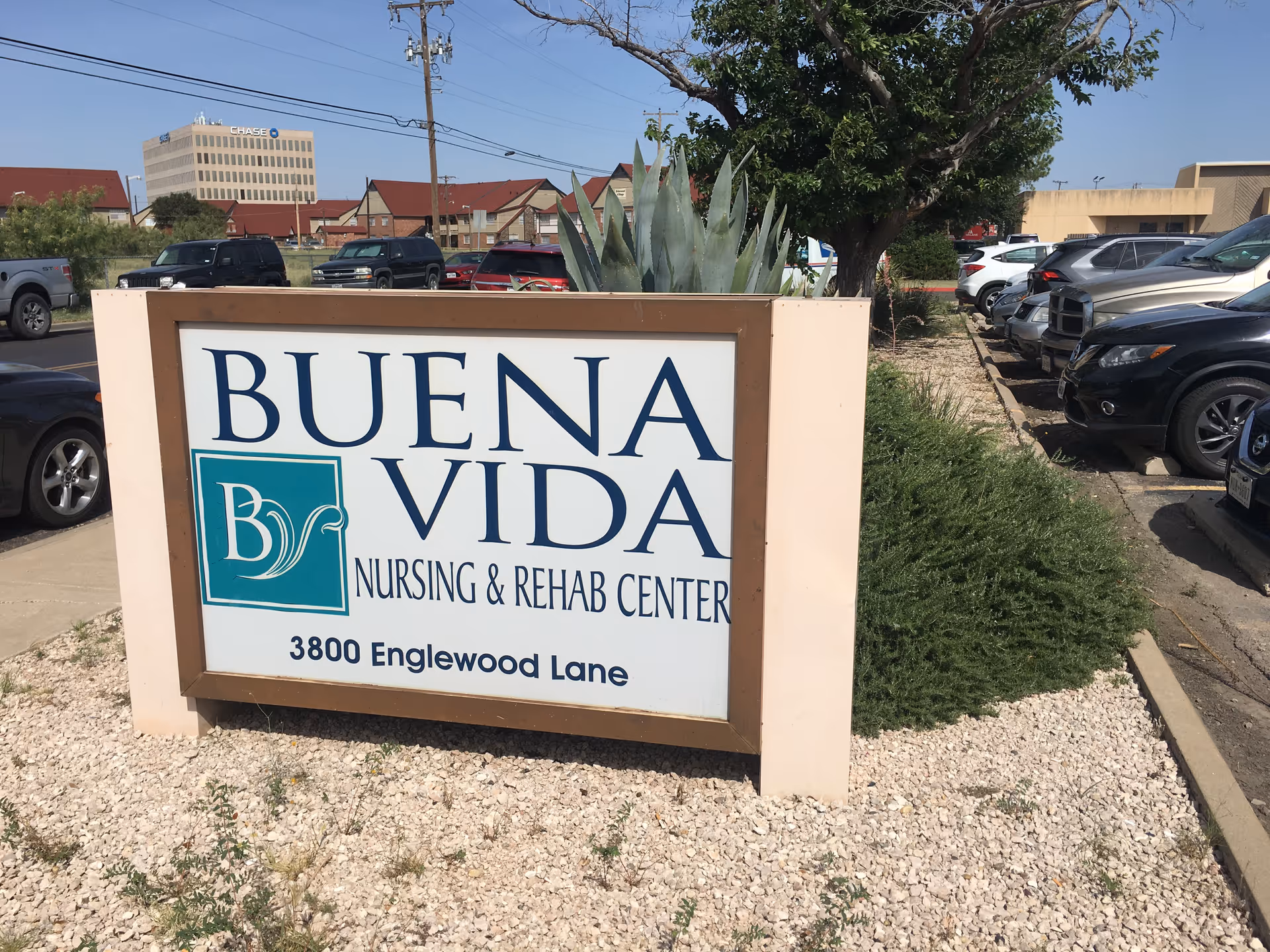 Outdoor view of a sign for Buena Vida Nursing & Rehab Center located at 3800 Englewood Lane, surrounded by gravel and greenery with parked cars and buildings in the background under a clear sky.