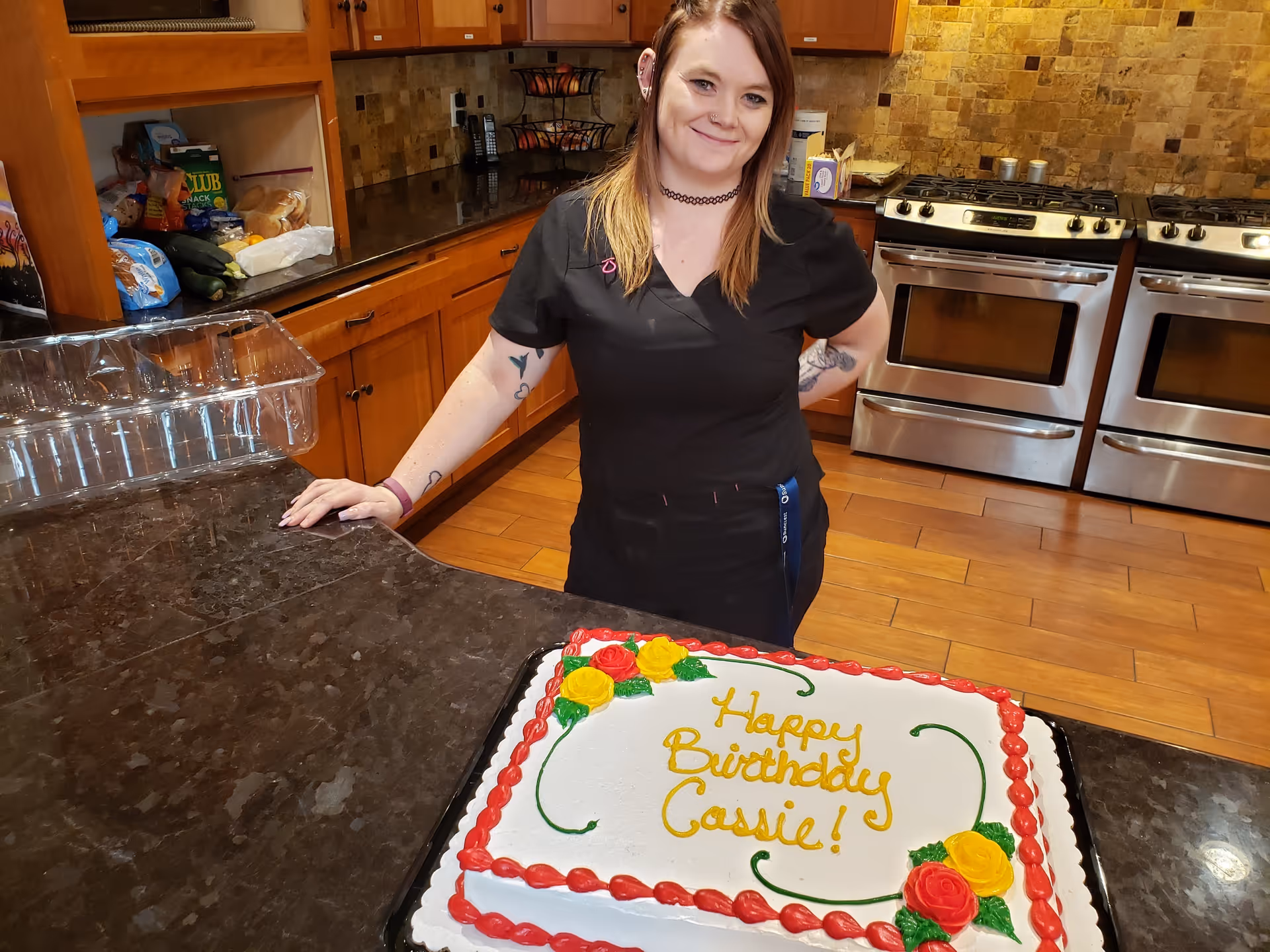 A woman with tattoos and a black outfit stands behind a kitchen counter with a birthday cake that says 'Happy Birthday Cassie!'. The kitchen has wooden cabinets, a tiled backsplash, and two stainless steel ovens.