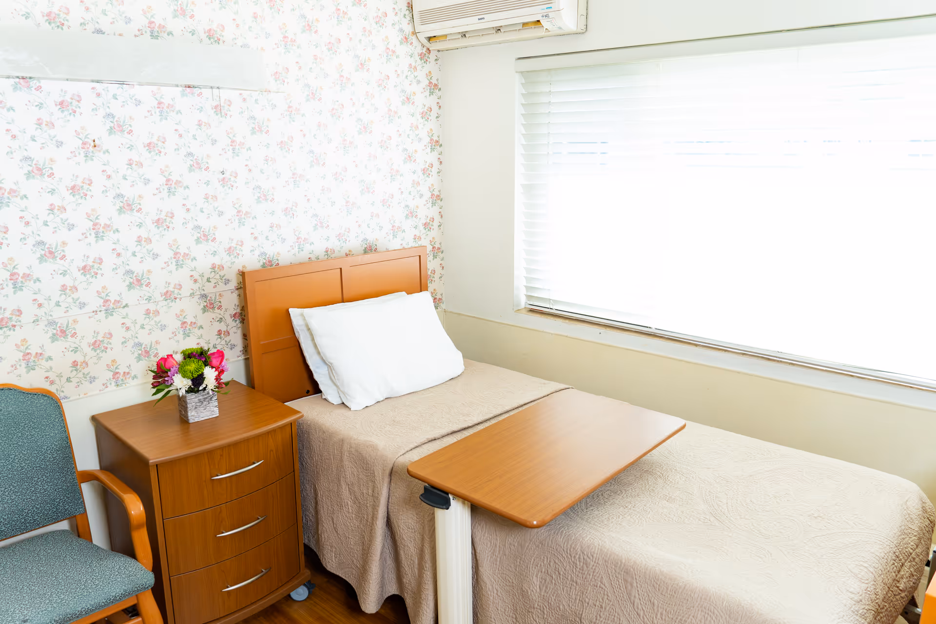 A bright single-occupancy bedroom with a twin bed, overbed table, wooden nightstand with flowers, a chair, floral wallpaper and a large window with blinds.