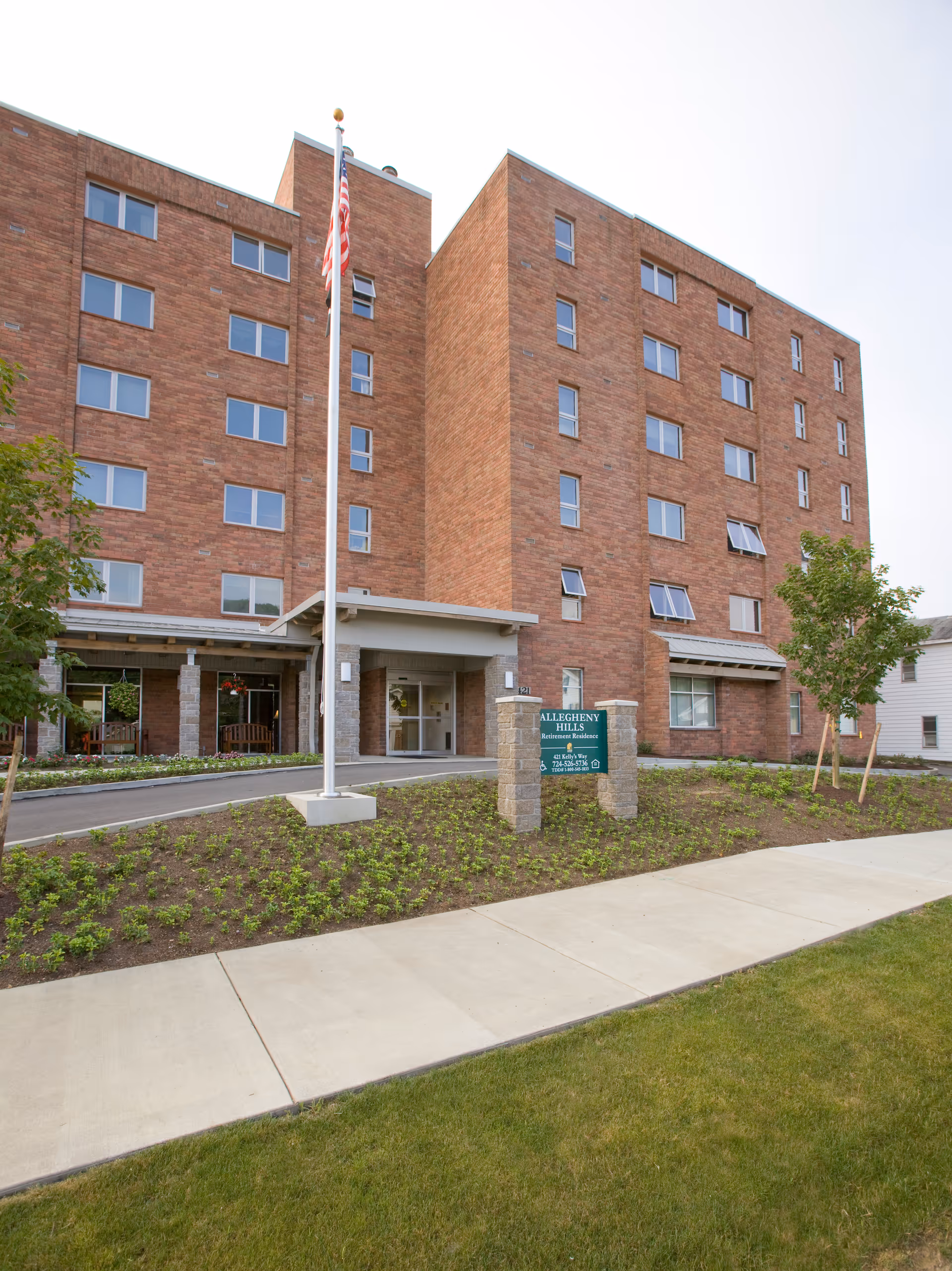 Brick multi-story retirement residence building with a covered entrance, flagpole, landscaping, and a sign reading 'Allegheny Hills'.