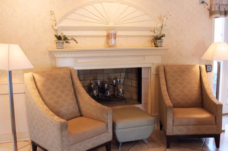 A cozy seating area with two patterned armchairs and a small ottoman placed in front of a decorative fireplace. The fireplace mantel is adorned with two potted plants and a glass vase. Two floor lamps with white shades flank the chairs, and a window with curtains is visible on the right side.