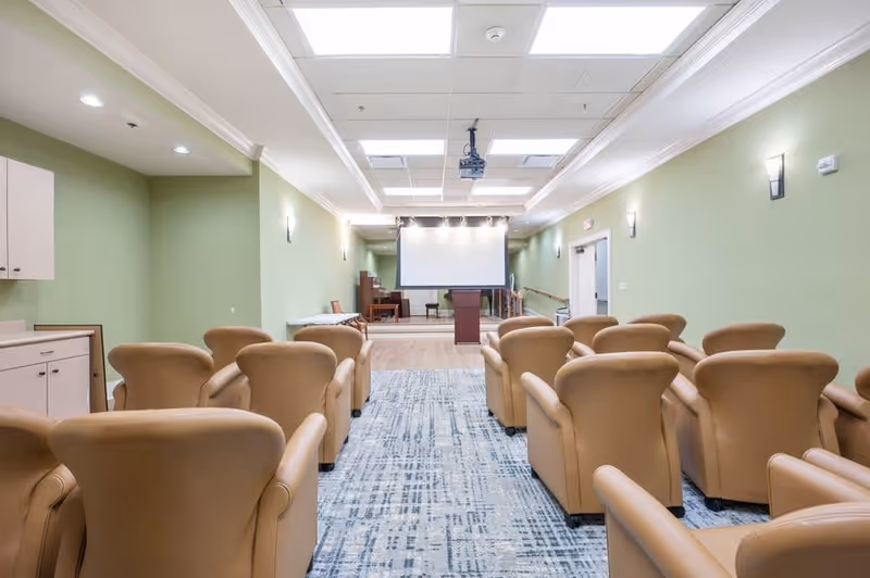 A small auditorium or meeting room with beige armchairs arranged in rows facing a projector screen and podium. The room has light green walls, a patterned carpet, ceiling lights, and a piano in the back corner.