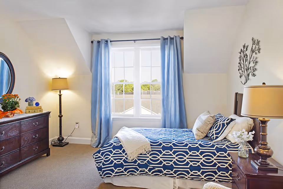 A bright bedroom with a single bed covered in a blue and white patterned bedspread and pillows. The room features a large window with light blue curtains, a wooden dresser with a round mirror, a floor lamp, and a bedside table with a lamp and a small vase of flowers. The walls are light-colored and decorated with a metal wall art piece.