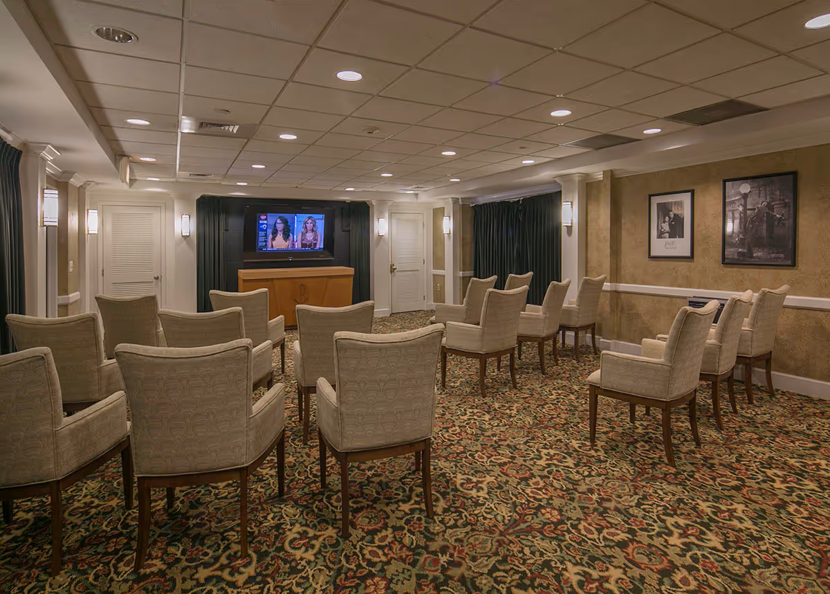 A small theater or media room with multiple beige upholstered chairs arranged in rows facing a flat-screen TV mounted on a wooden cabinet. The room has patterned carpet, beige walls with framed pictures, and recessed ceiling lights.