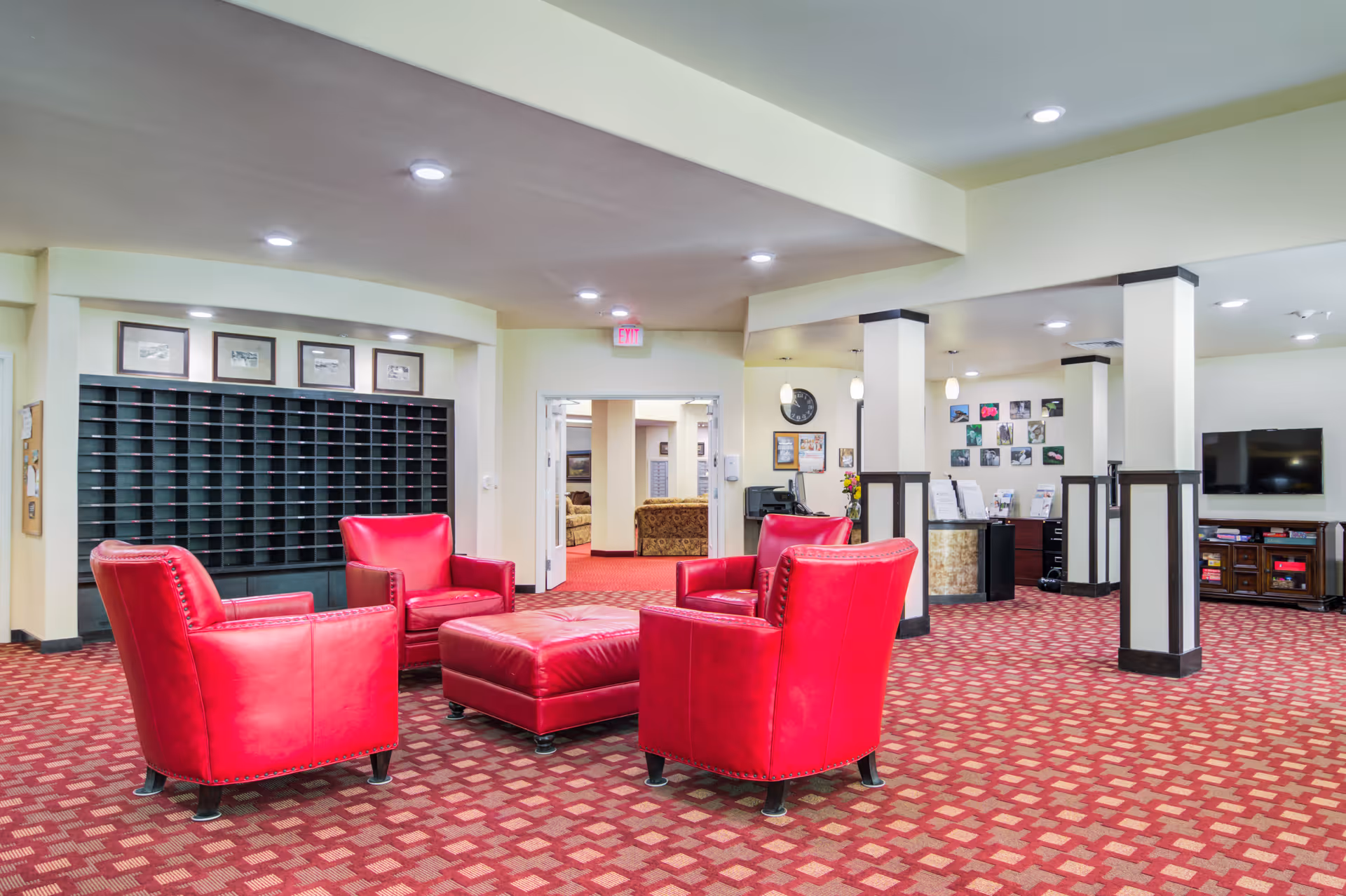 A spacious interior common area with four red leather armchairs arranged around a matching red leather ottoman on a patterned red carpet. The room has white walls with black trim, recessed lighting, and several columns. In the background, there is a reception desk, a wall-mounted TV, and a mail cubby unit with framed pictures above it.