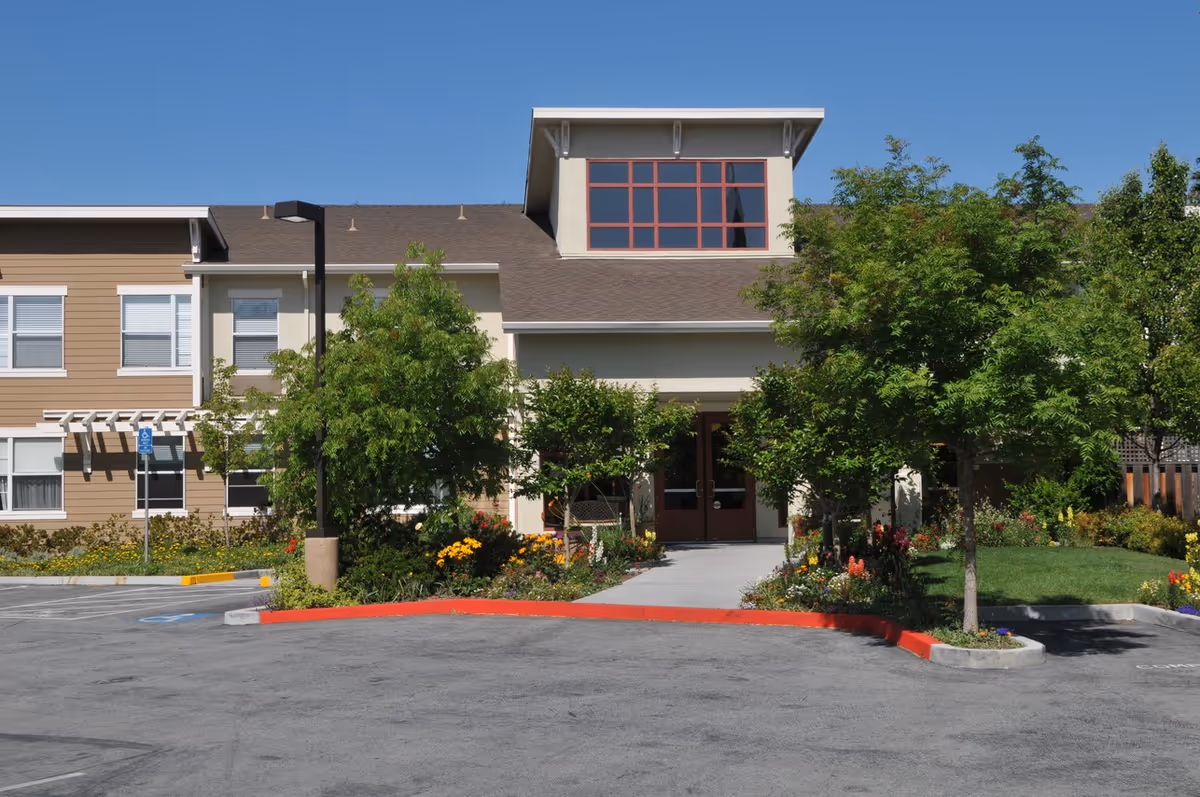 Front exterior view of a senior living facility named Pacific Gardens with a clear blue sky, trees, flowers, and a paved driveway leading to the entrance.